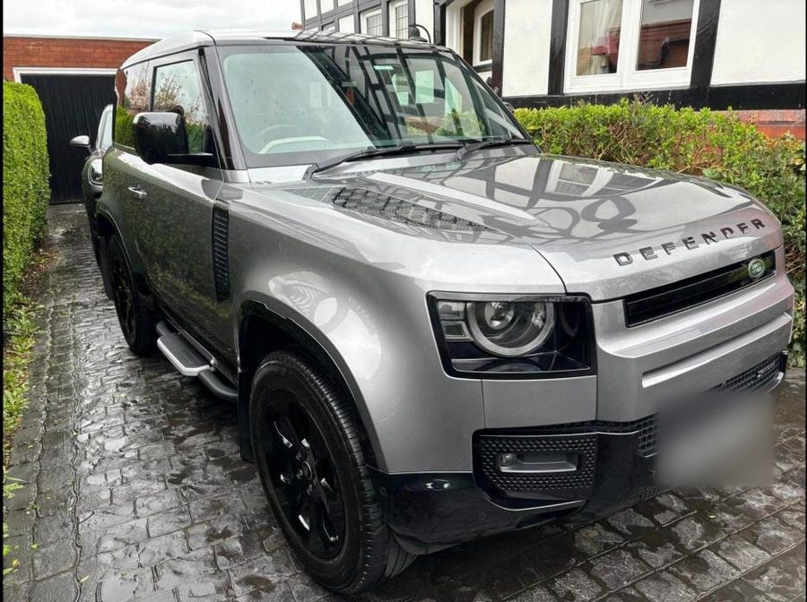 Silver-grey Land Rover Defender parked on a wet block-paved drive outside a house.