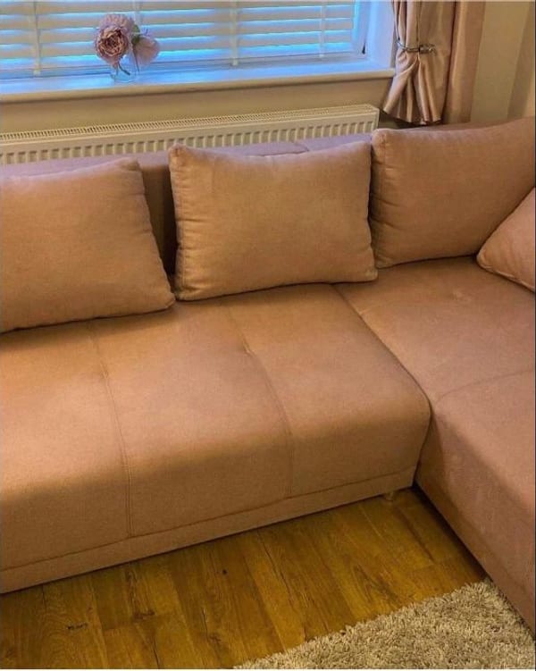 Blush pink corner sofa with cushions, under a window with white blinds.